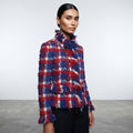A model wearing a red, blue, and white fringed tweed plaid jacket with black trousers, standing against a light gray studio background.